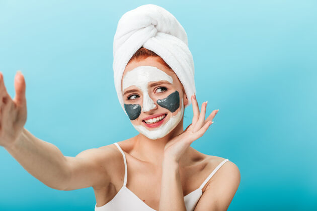 宠爱带着面具的微笑女人在自拍蓝色背景下 一个头上戴着毛巾的快乐女人的摄影棚照片大笑擦洗治疗