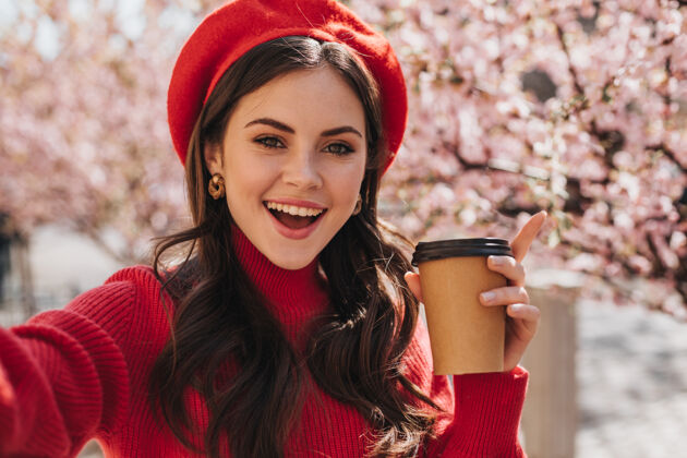 女性红衣美女捧着一杯茶 在樱花的背景下自拍黑发女孩戴着帽子微笑 拿着咖啡杯摆姿势的肖像开花华丽生活