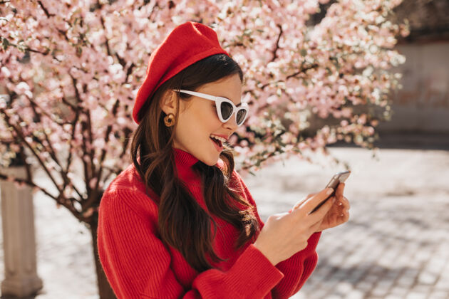温柔戴着贝雷帽和太阳镜的美女在樱花附近打电话聊天外面是一张穿着红色现金棉毛衣拿着手机的女士的画像春天天芳香