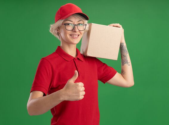 眼镜年轻的送货员身穿红色制服 戴着帽子 戴着眼镜 肩上扛着纸板箱 微笑着自信地竖起大拇指站在绿色的墙上肩膀交货拇指