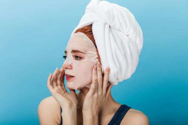 女性梦幻女孩在蓝色背景上涂上隔离的面膜摄影棚拍摄的头上戴着毛巾的年轻女士移开视线护理人面部