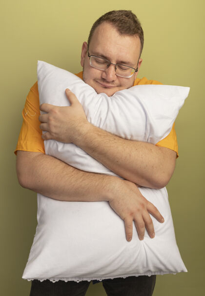 感觉戴着眼镜的男人穿着橘色衬衫抱着枕头闭着眼睛站在光墙上感受积极的情绪情绪穿枕头