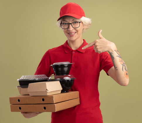 指着身穿红色制服 戴着眼镜 手持比萨饼盒和食品包的年轻送货员站在绿色的墙上 用食指指着他们 开心地微笑着眼镜制服帽子