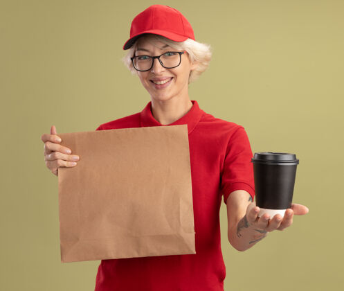 眼镜身穿红色制服 戴着帽子 戴着眼镜 拿着纸包 端着咖啡杯 微笑着站在绿色的墙上帽子咖啡提供