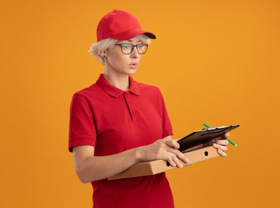 帽子身穿红色制服 戴着帽子 戴着眼镜 拿着披萨盒和铅笔的剪贴板的年轻送货员站在橙色的墙上 神情迷茫 非常焦虑焦虑拿着眼镜