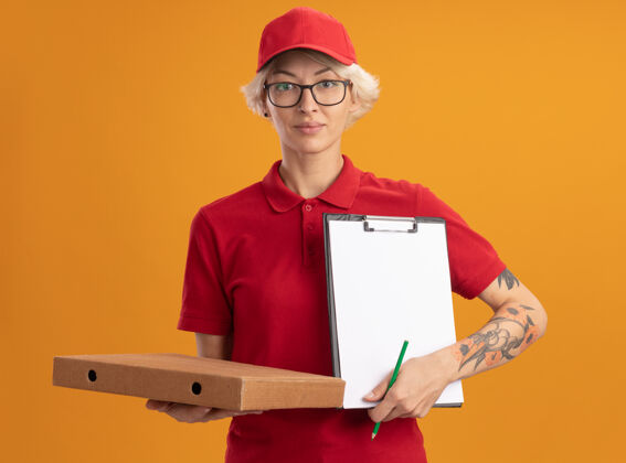 眼镜身穿红色制服 戴着眼镜 拿着披萨盒和铅笔的剪贴板的年轻送货员自信地站在橙色的墙上帽子拿着制服