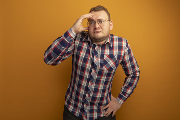 头一个戴着眼镜和格子衬衫的男人站在橙色的墙上 困惑地看着一边 手放在头上眼镜手靠边