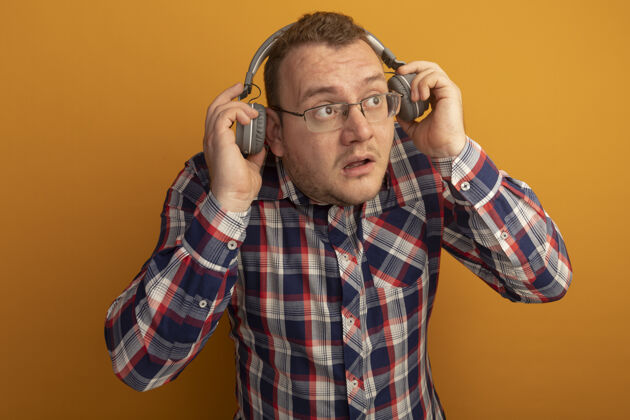 男人一个戴着眼镜和格子衬衫戴着耳机的男人站在橙色的墙上 站在一旁困惑地看着耳机眼镜站着