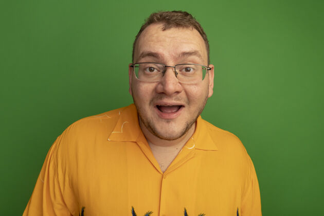 眼镜戴着眼镜和橘色衬衫的快乐男人站在绿色的墙上惊讶地笑了惊喜站起来伙计