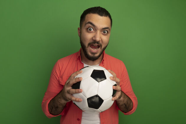 抱着情绪激动的留着胡子的年轻人穿着橘色衬衫 手里拿着足球 站在绿色的墙上大声喊叫年轻人情绪球