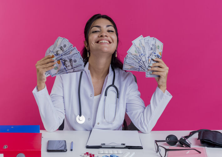听诊器快乐的年轻女医生穿着医用长袍和听诊器坐在办公桌旁 手里拿着医疗工具 手里拿着隔离在粉红色墙上的钱长袍拿着看