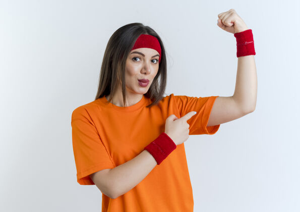 年轻自信的年轻运动女性戴着头带和腕带做着强烈的手势 指着她隔离在白色墙壁上的肌肉复制空间手势腕带佩戴