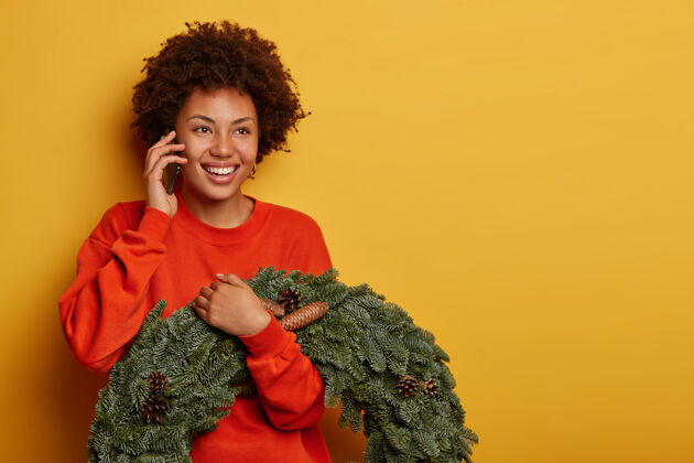 传统快乐的卷发女人喜欢电话交谈 和朋友讨论圣诞节的准备 手持松果的杉木手工花环 站在黄色的背景下季节花圈快乐