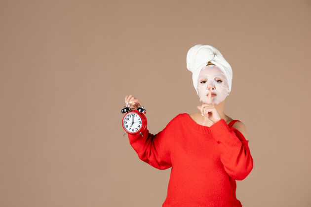 抱着正面图：戴着面具的年轻女性 手持粉色背景上的时钟安静护肤脸