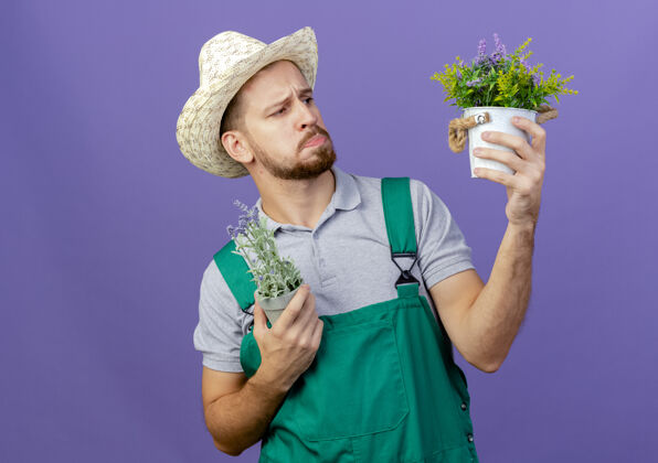 斯拉夫体贴的年轻英俊的斯拉夫园丁 穿着制服 戴着帽子 看着紫色墙壁上孤立的花盆和复制空间思想花园拿着