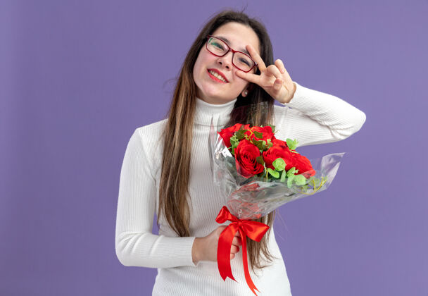玫瑰穿着休闲服的年轻漂亮女人手持一束红玫瑰看着镜头快乐而积极地展示着v字情人节的概念站在紫色的墙上举行天年轻