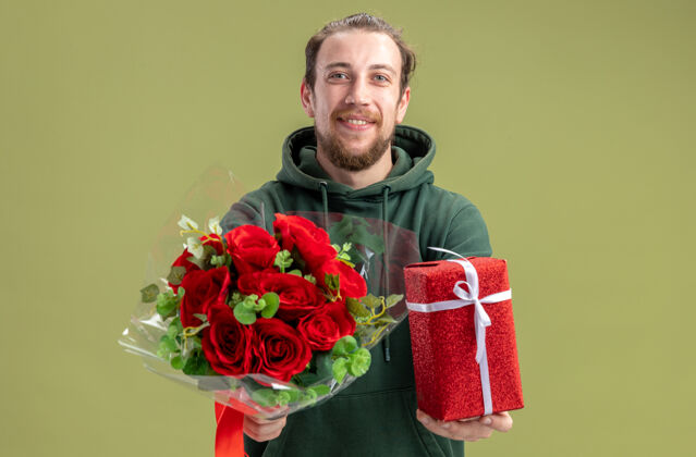 年轻快乐的年轻人穿着休闲服 带着一束红玫瑰 给女友礼物 看着相机 微笑着欢快地站在绿色的墙边 情人节的概念情人节女朋友休闲