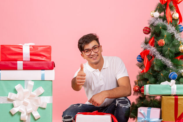 微笑前视图年轻人周围的礼物和圣诞树上的粉红色背景风景圣诞节礼物
