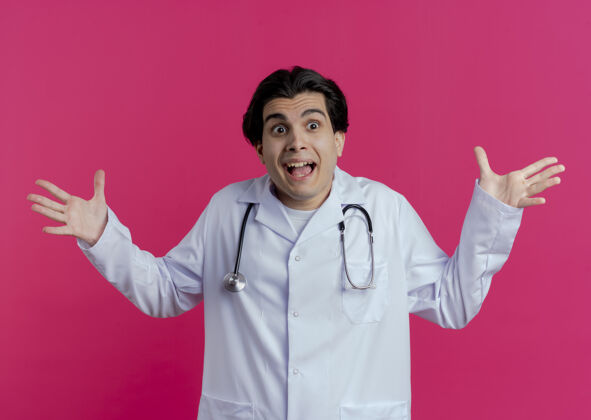 显示令人印象深刻的年轻男医生穿着医用长袍和听诊器看着侧面显示空手隔离在粉红色的墙上手男性长袍