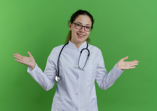 年轻快乐的年轻女医生穿着医用长袍 戴着听诊器 戴着眼镜 双手空空的隔离在绿色的墙上欢乐医疗表演