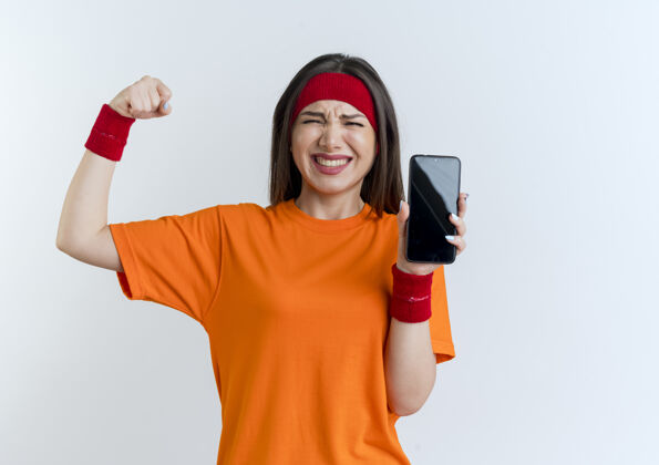 手势快乐的年轻运动女性戴着头带和腕带 显示手机做着强烈的手势 隔离在白色墙壁上 留有复制空间女士欢乐佩戴