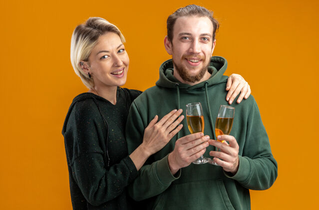 女人一对穿着休闲服 戴着香槟酒杯的快乐年轻夫妇站在橘色的墙上 脸上洋溢着欢笑庆祝情人节的快乐女人香槟看庆祝