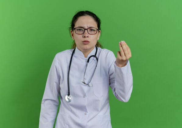 年轻人年轻的女医生穿着医用长袍 戴着听诊器 戴着眼镜 在绿色的墙上做着钱的手势 还有复印空间表情医生医疗