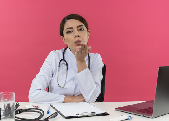 吹自信的年轻女医生穿着医用长袍 手持听诊器坐在办公桌旁 手里拿着医疗工具女性看信心