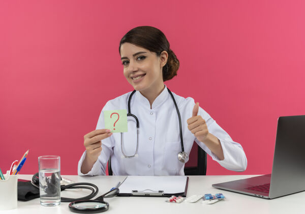 听诊器微笑着的年轻女医生穿着医用长袍和听诊器坐在办公桌旁 手里拿着医疗工具和手提电脑 手里拿着问号 拇指向上竖立在粉红色的墙上年轻人长袍微笑