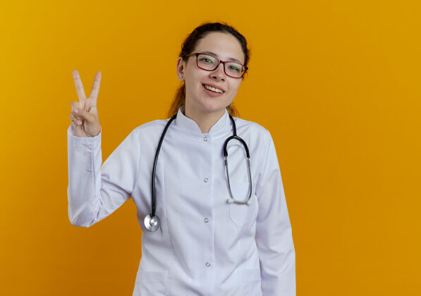 穿面带微笑的年轻女医生 穿着医用长袍 戴着听诊器 戴着一副眼镜 在橙色的墙上显示出一种与世隔绝的和平姿态医学医生眼镜