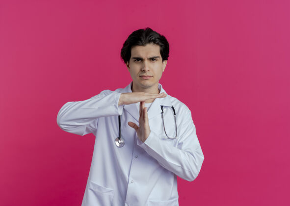 皱眉皱眉的年轻男医生穿着医用长袍和听诊器做超时手势隔离在粉红色的墙上与复制空间男性穿着表情