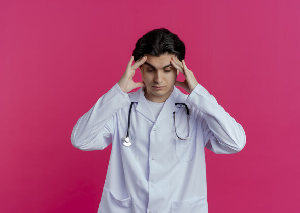 疲倦疲惫的年轻男医生穿着医用长袍和听诊器 闭着眼睛摸着头 隔离在粉红色的墙上 留着复印空间封闭医疗长袍