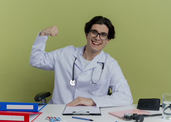 医生快乐的年轻男医生穿着医用长袍 戴着听诊器 戴着眼镜 坐在办公桌旁 用医疗工具做着强烈的手势 隔离在橄榄绿的墙上眼镜强壮办公桌