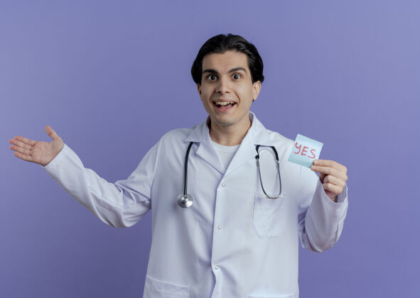 空令人印象深刻的年轻男医生穿着医用长袍和听诊器显示是的笔记和空手隔离在紫色的墙上听诊器显示年轻