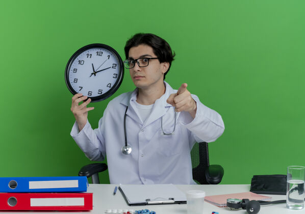 印象年轻的男医生穿着医用长袍 戴着听诊器 戴着眼镜 坐在办公桌旁 拿着医疗工具 拿着时钟 看着绿色的墙上孤立的指针钟表指着医生