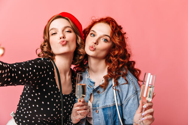 卷发两个女孩拿着酒杯自拍朋友们在粉色背景下喝香槟的摄影棚照片法国欢乐派对