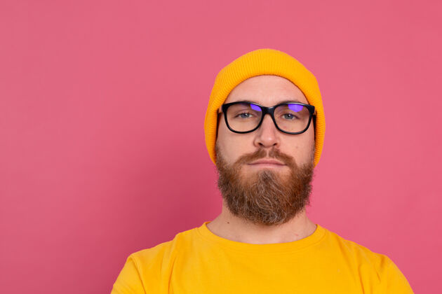 帅气时尚帅气的留着胡子的欧洲男人的肖像 穿着休闲的黄色衬衫 戴着粉红色的帽子和眼镜正面思考时尚