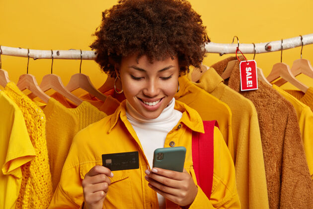 年轻人皮肤黝黑的女人使用现代手机和信用卡 网上购物 通过互联网下单 插入银行账户信息 靠着衣架站立销售包购物