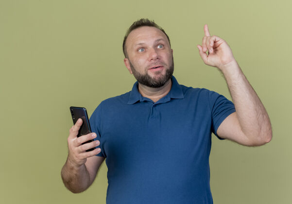 提高令人印象深刻的成年斯拉夫男子手持手机 一边看着一边举起手指橄榄色成人斯拉夫