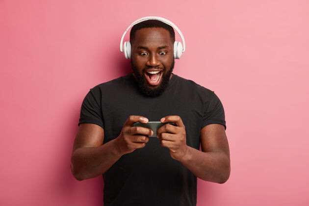 玩兴奋的大胡子男人横拿着现代智能手机 玩网络游戏 使用heeadset 贴在小玩意屏幕上 穿着黑色t恤 一个人玩得很开心非洲裔安装兴奋