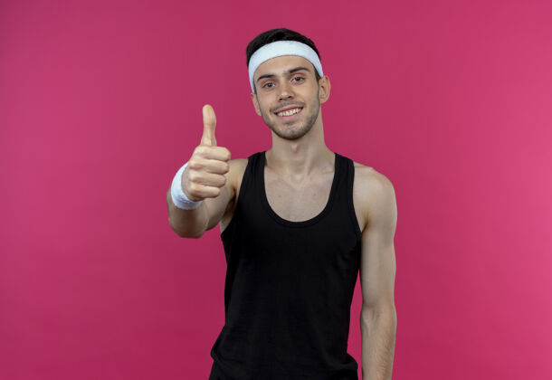 积极戴着头巾的年轻的运动型男人微笑着快乐而积极地向粉色竖起大拇指看向上粉红