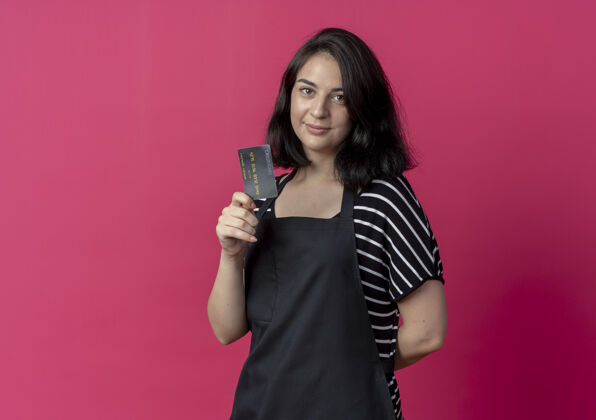 信用卡穿着围裙的年轻漂亮的女理发师在镜头前展示信用卡的样子 脸上露出粉红色的微笑看漂亮微笑