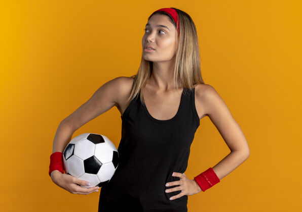 年轻身穿黑色运动服 头戴红色头带 手持足球 神情自信地看着橙色的年轻健身女孩健身足球橙色