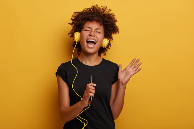 感觉情绪化的黑皮肤黑发女人举起手臂 高声唱歌 听音乐 带来愉快的回忆隔离在黄色耳机科技旋律
