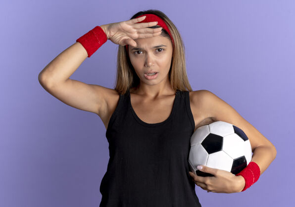年轻身穿黑色运动服 头戴红色头带 手持足球的年轻健身女孩 手举蓝色头顶 遥望远方黑色看球