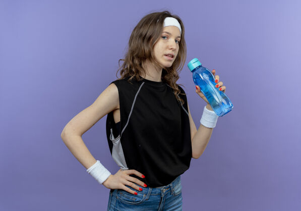头带穿着黑色运动服的年轻健身女孩 戴着头巾 手里拿着一瓶水 严肃的脸盖着蓝色健身年轻脸