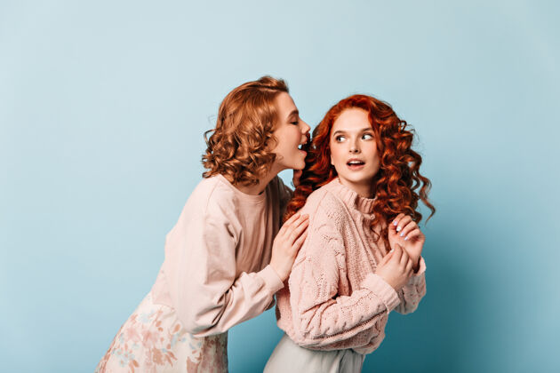 两个人卷发女孩在蓝色背景上交谈摄影棚拍摄的穿着时髦衣服的女性朋友时尚在一起交谈