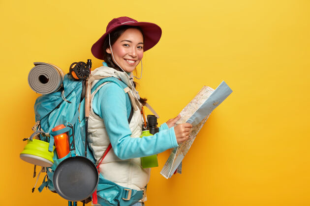 活动活跃的韩国女游客背着大背包 戴着帽子和休闲服 拿着地图 学习路线 有很多旅行中需要的东西游客姿势微笑