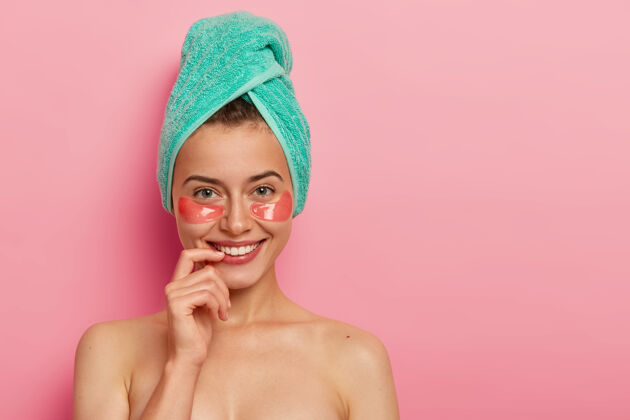 裸体高兴的欧洲女人照顾眼睛周围娇嫩的皮肤 贴胶原蛋白贴片 化最少的妆 头上裹着浴巾 赤裸着站在粉色的背景下路径美容化妆品