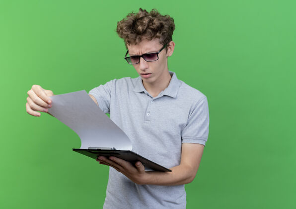 衬衫戴着黑眼镜的年轻人 穿着灰色马球衫 手里拿着一张白纸的剪贴板 严肃地看着它 脸上泛着绿色严重马球绿色